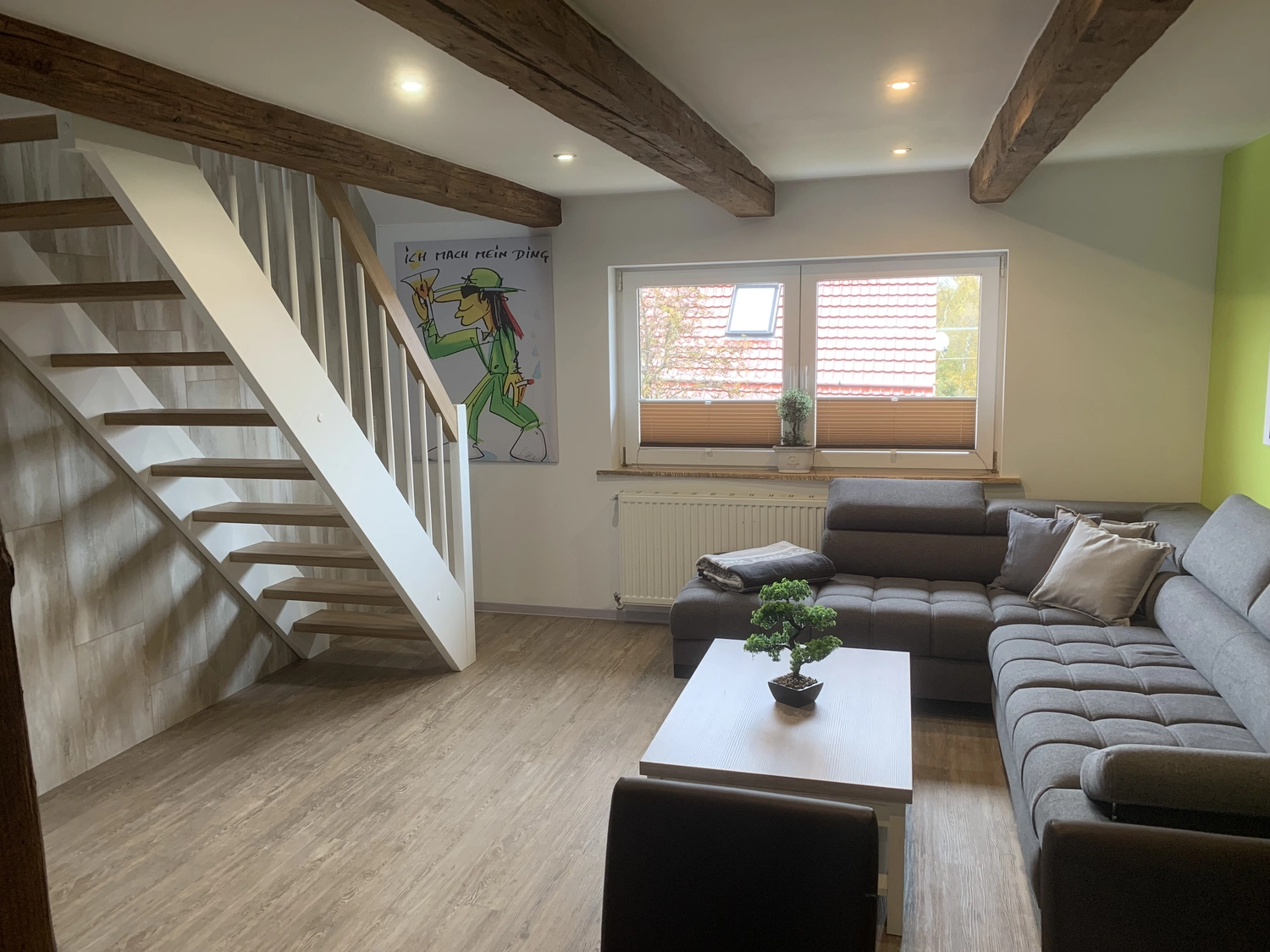 Ecklebe - Ferienwohnung - Ein Wohnzimmer mit Holztreppe, grauem Ecksofa, Couchtisch und sichtbaren Deckenbalken.